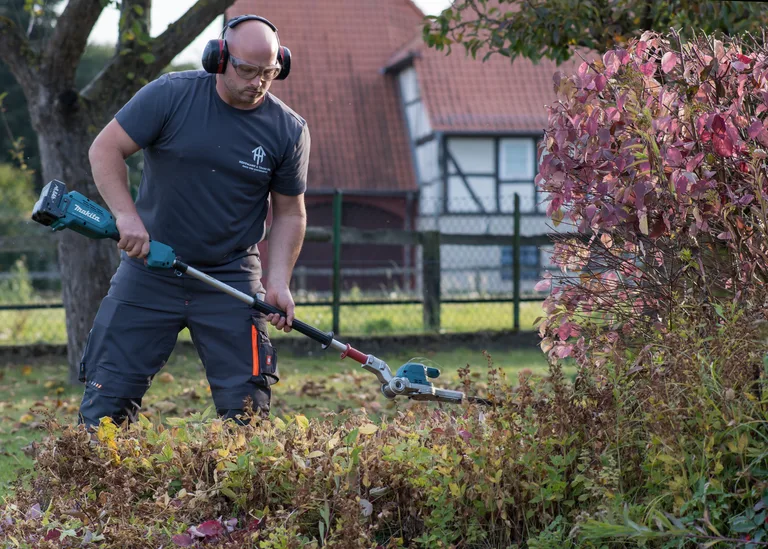 Einer der Inhaber schneidet eine niedrige Hecke mit einer langen Akku-Heckenschere